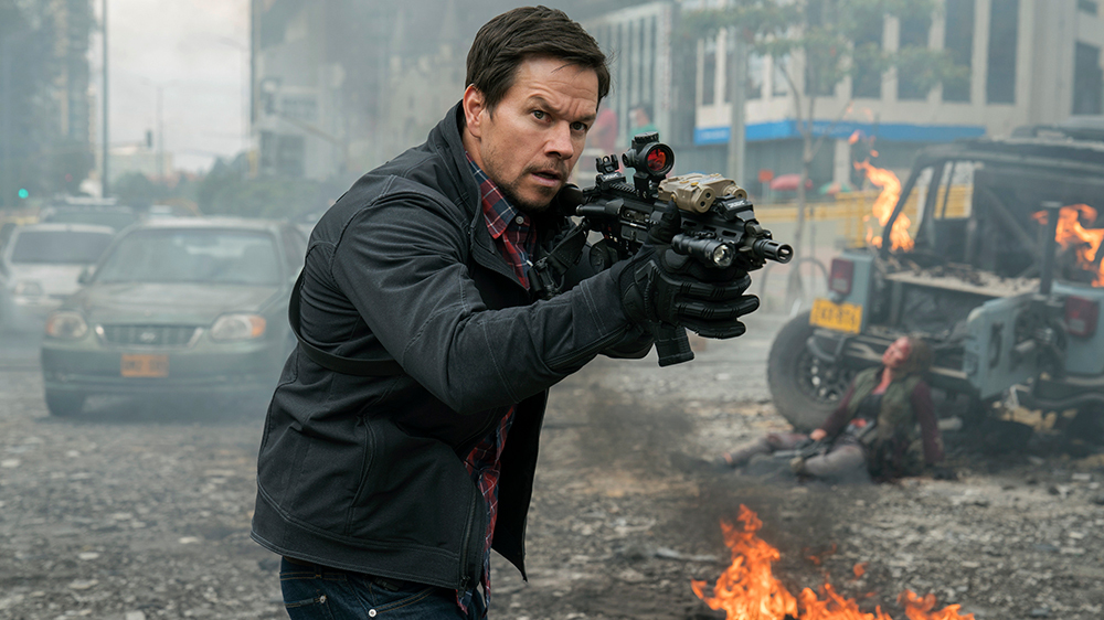 Mark Wahlberg points a gun in a tactical stance with burning cars behind him in Mile 22.