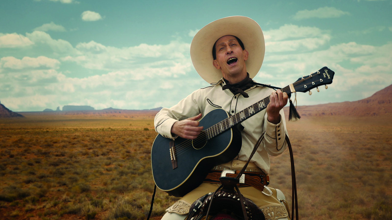 Tim Blake Nelson on a horse wearing white playing a guitar and singing in front of a picturesque landscape in the film The Ballad of Buster Scruggs