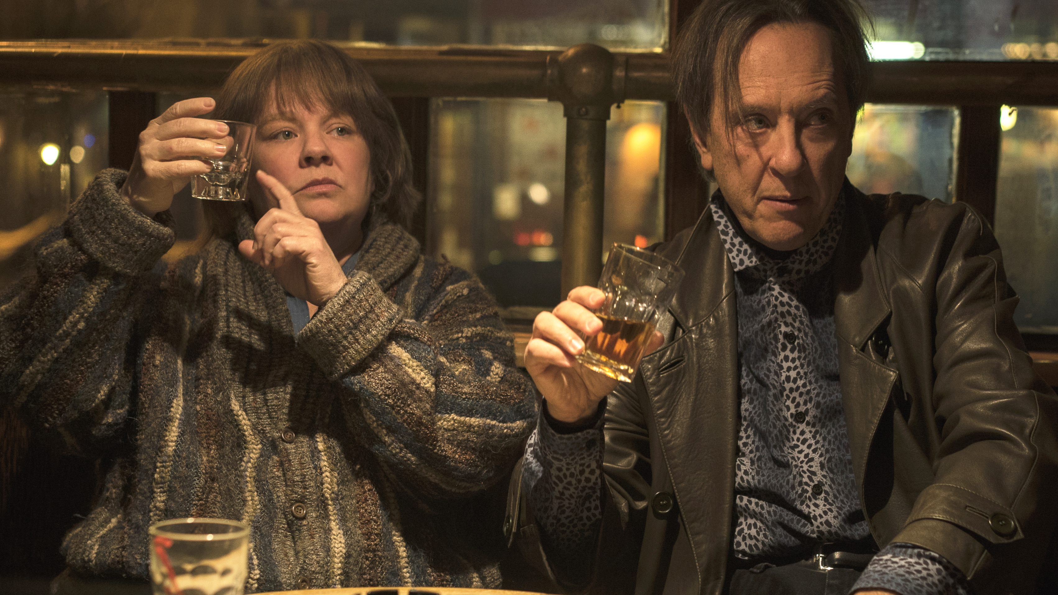 Melissa McCarthy and Richard E. Grant in the movie Can You Ever Forgive Me? are sitting at a bar holding up their glasses of liquor asking for more.