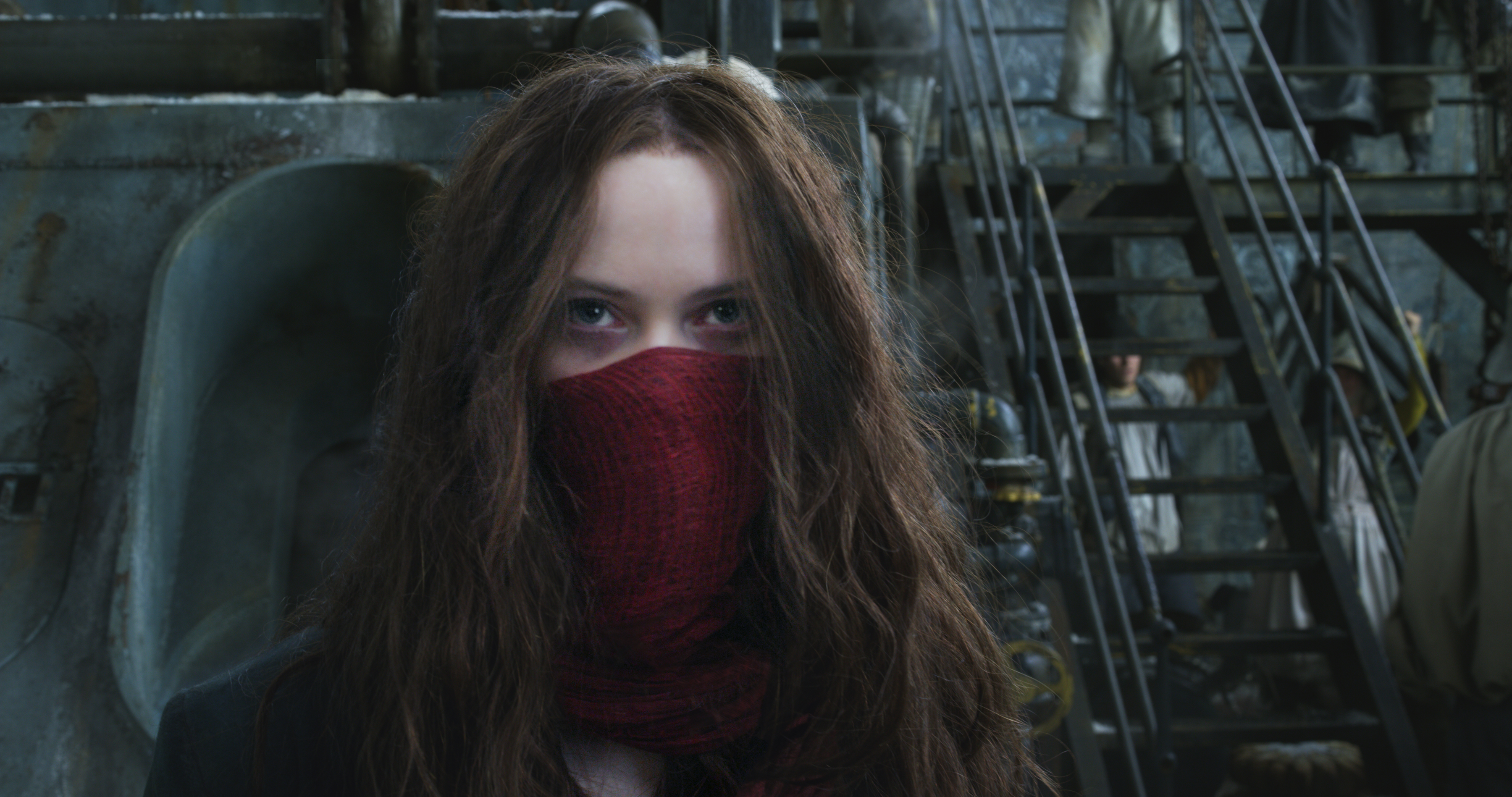 Hera Hilmar wearing a red scarf for a mask and looking intently in front of some stairs and machinery in the movie Mortal Engines
