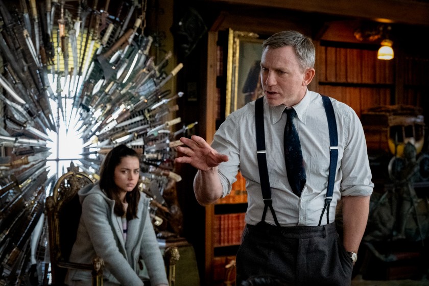 Daniel Craig gestures with his hand laying out his theory while Ana de Armas sits by a throne of knives in the movie Knives Out.