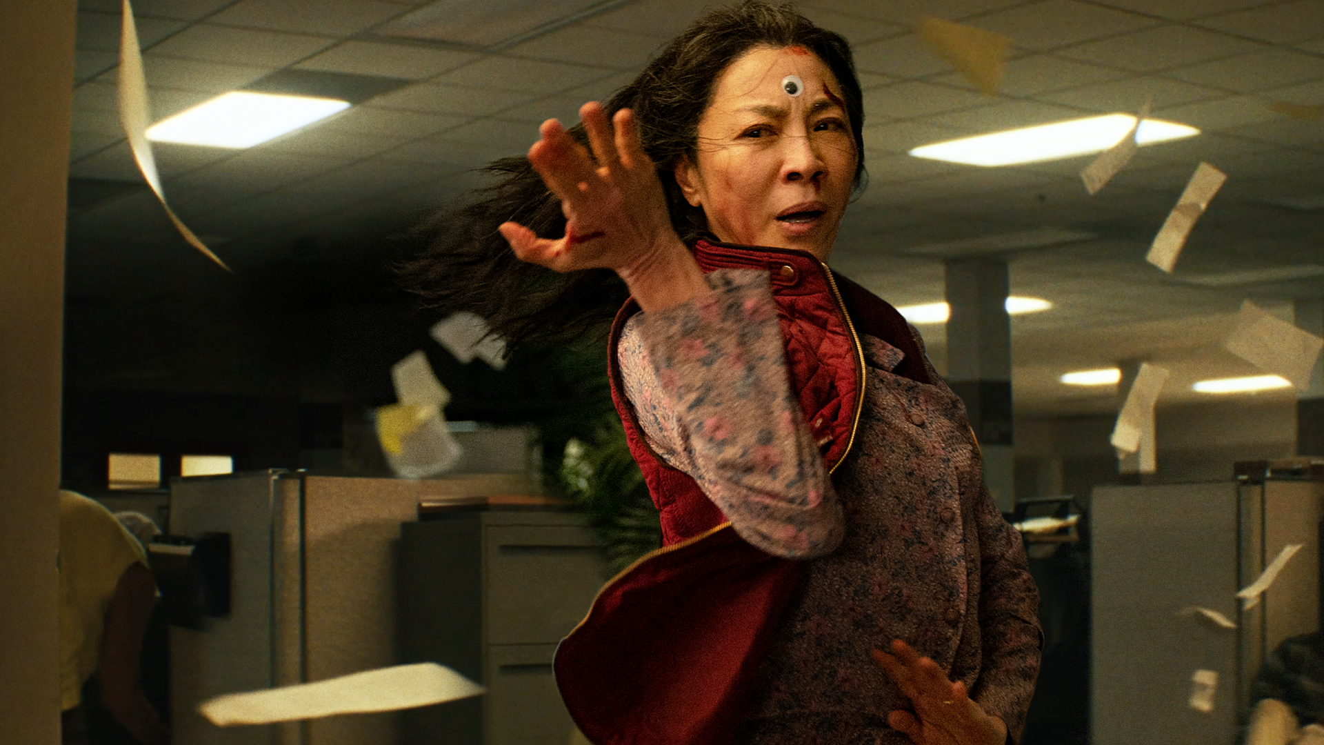 Michelle Yeoh sweeps her arm in a martial arts move with flying papers and a googly eye on her forehead in the movie Everything Everywhere All at Once