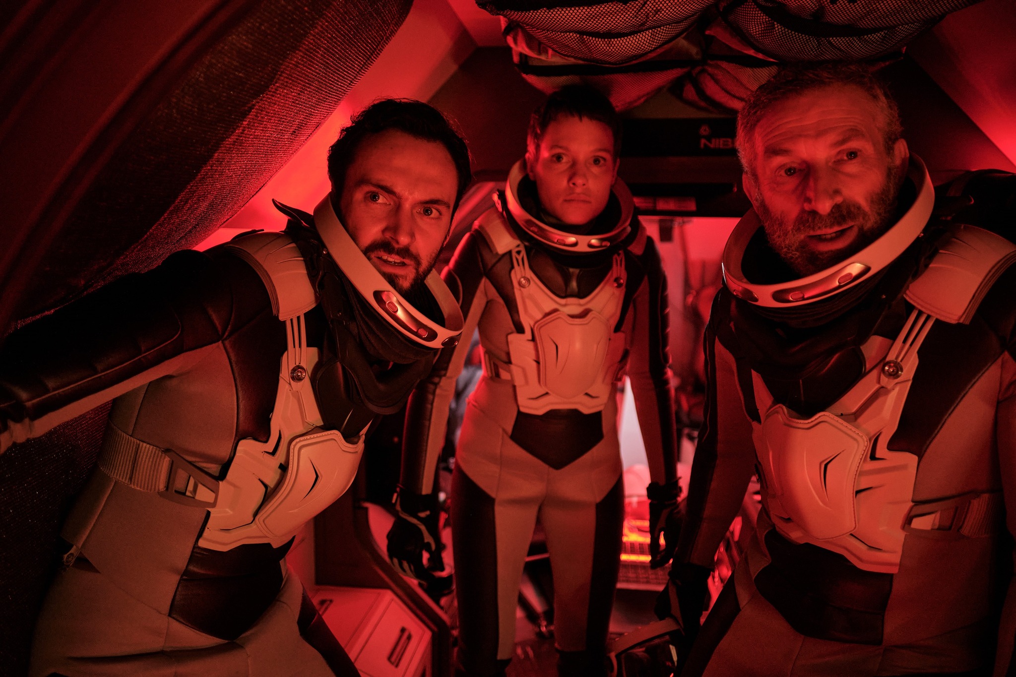 George Blagden, Julia Franz Richter, and Mark Ivanir stand in space suits bathed in red light looking ahead worriedly in the movie Rubikon