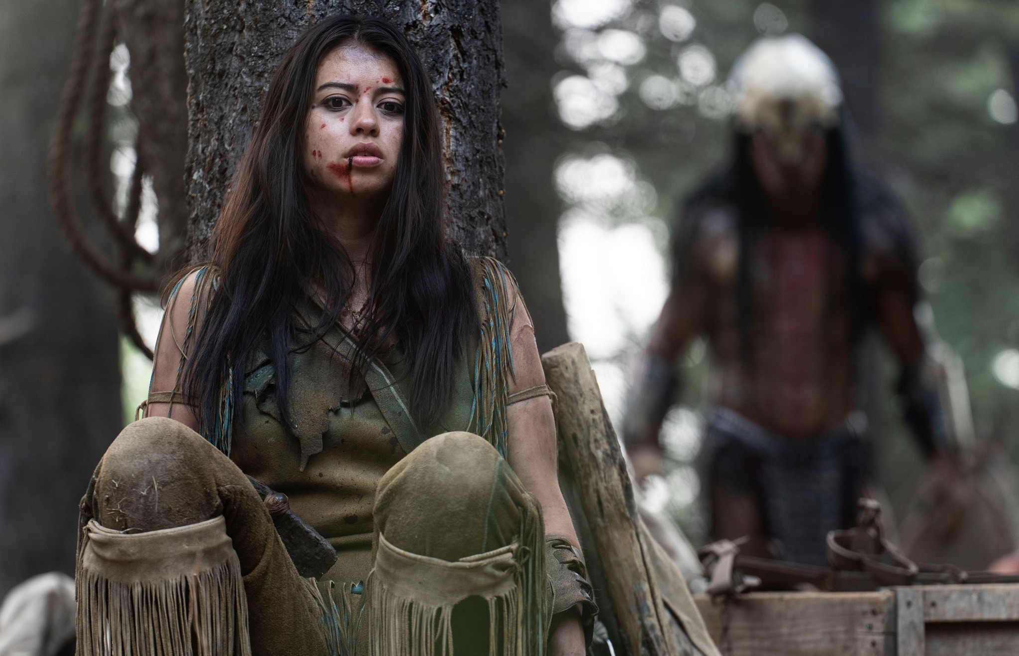 Amber Midthunder hides behind a tree while a Predator stalks her in the movie Prey
