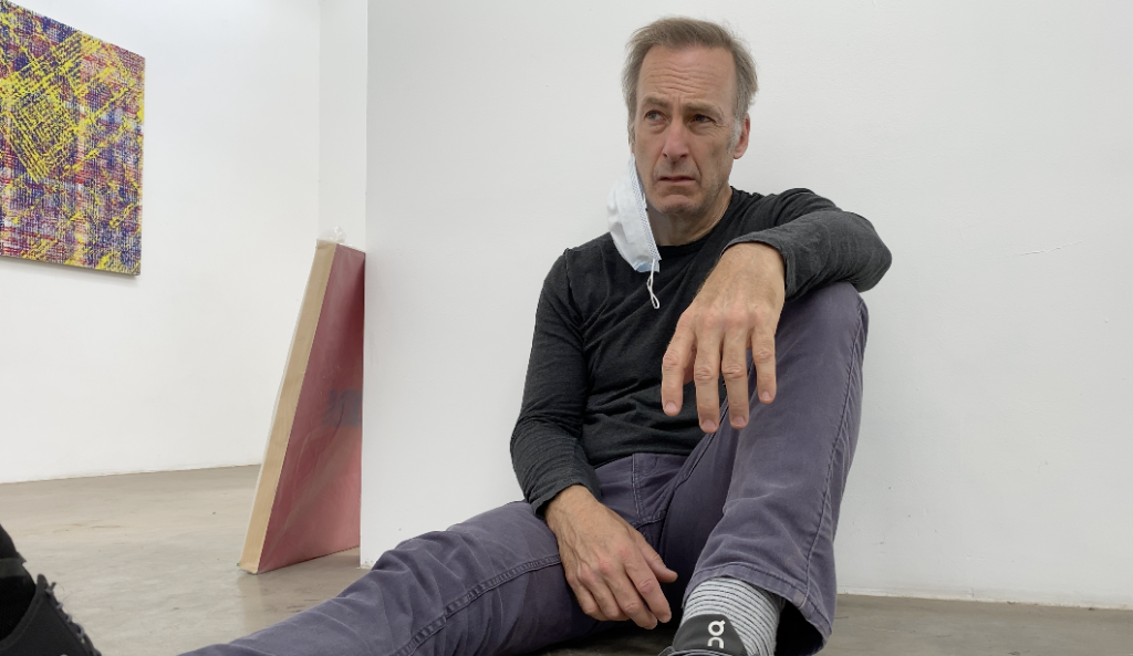 Bob Odenkirk sits against a wall looking miserable with a mask dangling from his face in the film Life Upside Down.