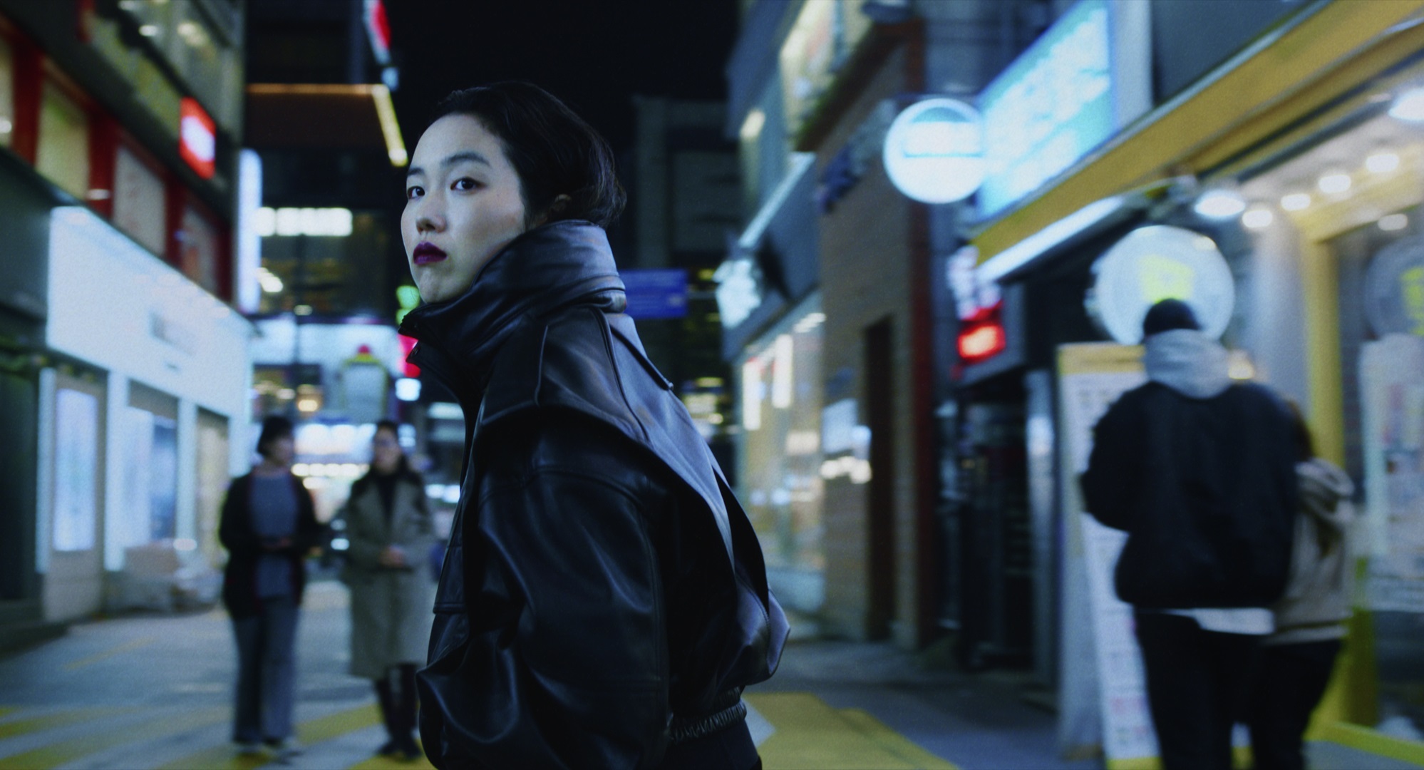 Park Ji-Min stands in a leather jacket on a street in Korea at night, looking back, in the movie Return to Seoul.